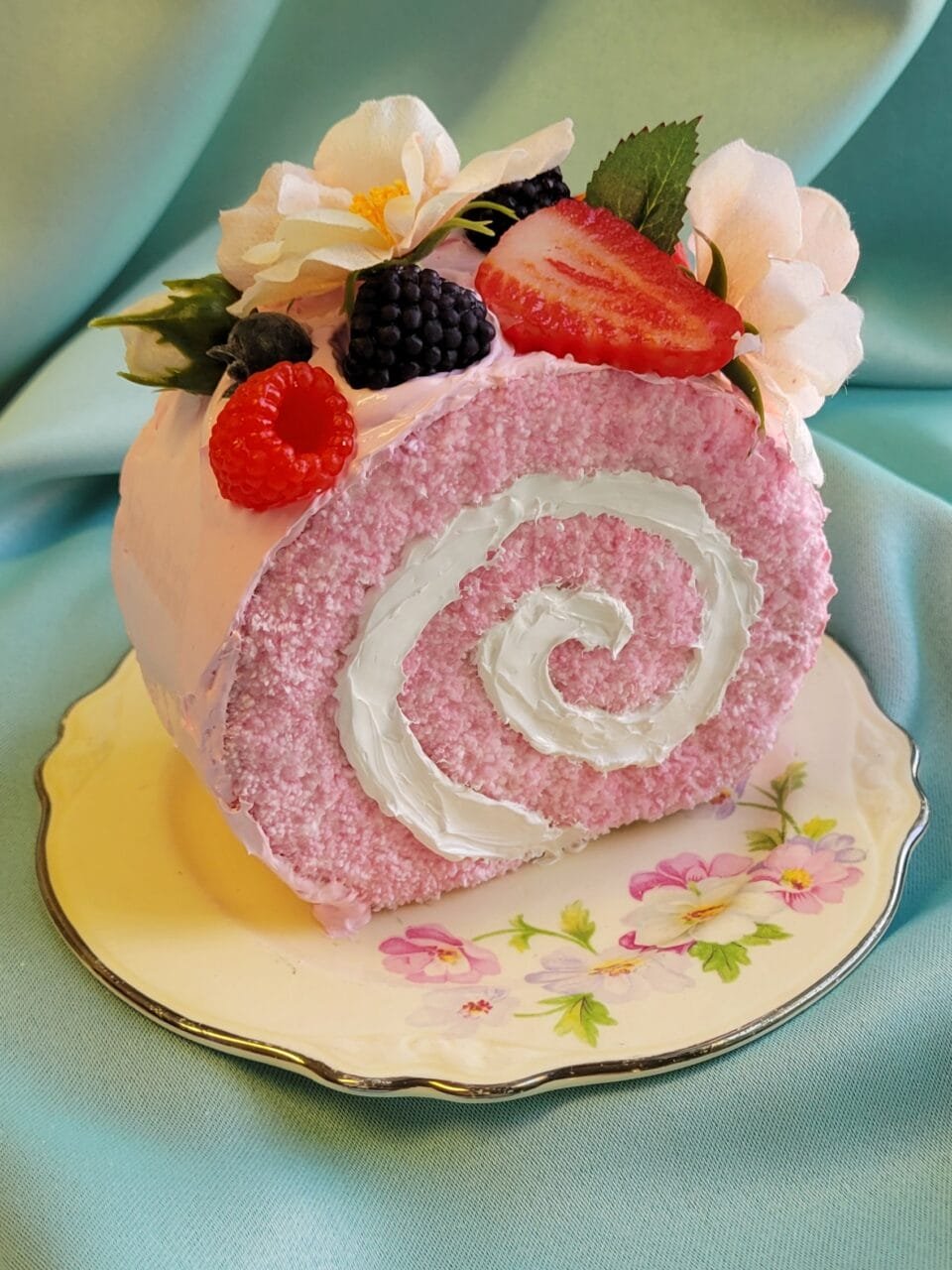 a small sculpture of a slice of a Swiss roll on an ornate plate, with realistic frosting and fruit details and embroidery that looks like the texture of fluffy sponge