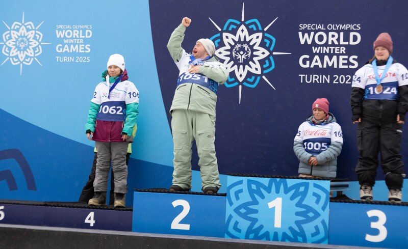 An athlete stands on a podium after finishing in second place in a snowboarding competition. His fellow athletes, in third and fourth place, stand with him