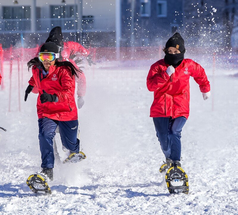 Two women in red jackets wearing snowshoes sprint through the snow.