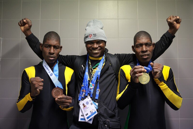 three men stand in celebration. On the outside, two twin brothers hold up their medals. Between them, a proud coach raises his hands and smiles.