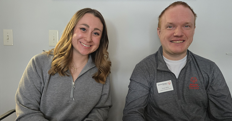 A woman (left) and man (right) sit in front of a grey wall. They both have on gray pullovers and are smiling for the camera.