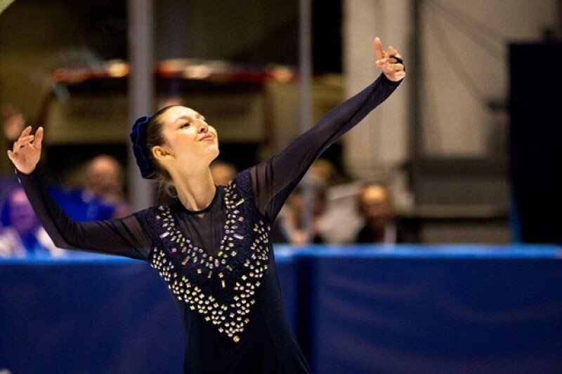 A figure skater is pictured performing a routine, her arms raised artistically. Blurred behind her are spectators.