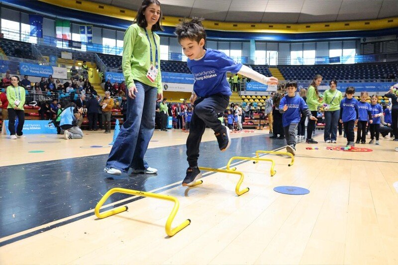 Young athletes jump over small hurdles in a course set up in a gymnasium.