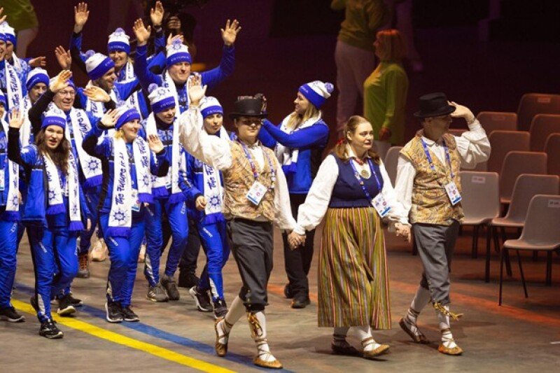 A group of three DanceSport athletes dressed in traditional Estonian costumes, walk into an arena. A group of Estonian athletes dressed in blue winter gear cheer from behind them