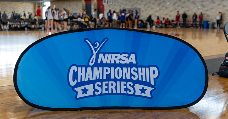 This photo shows a blue banner that says "NIRSA Championship Series" in white writing. Behind the banner, there is a basketball court with people on it.