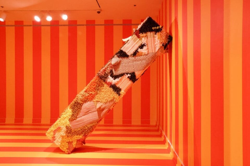 an installation view of a gallery with a leaning textile column-like sculpture, with the walls painted in wide orange and red stripes